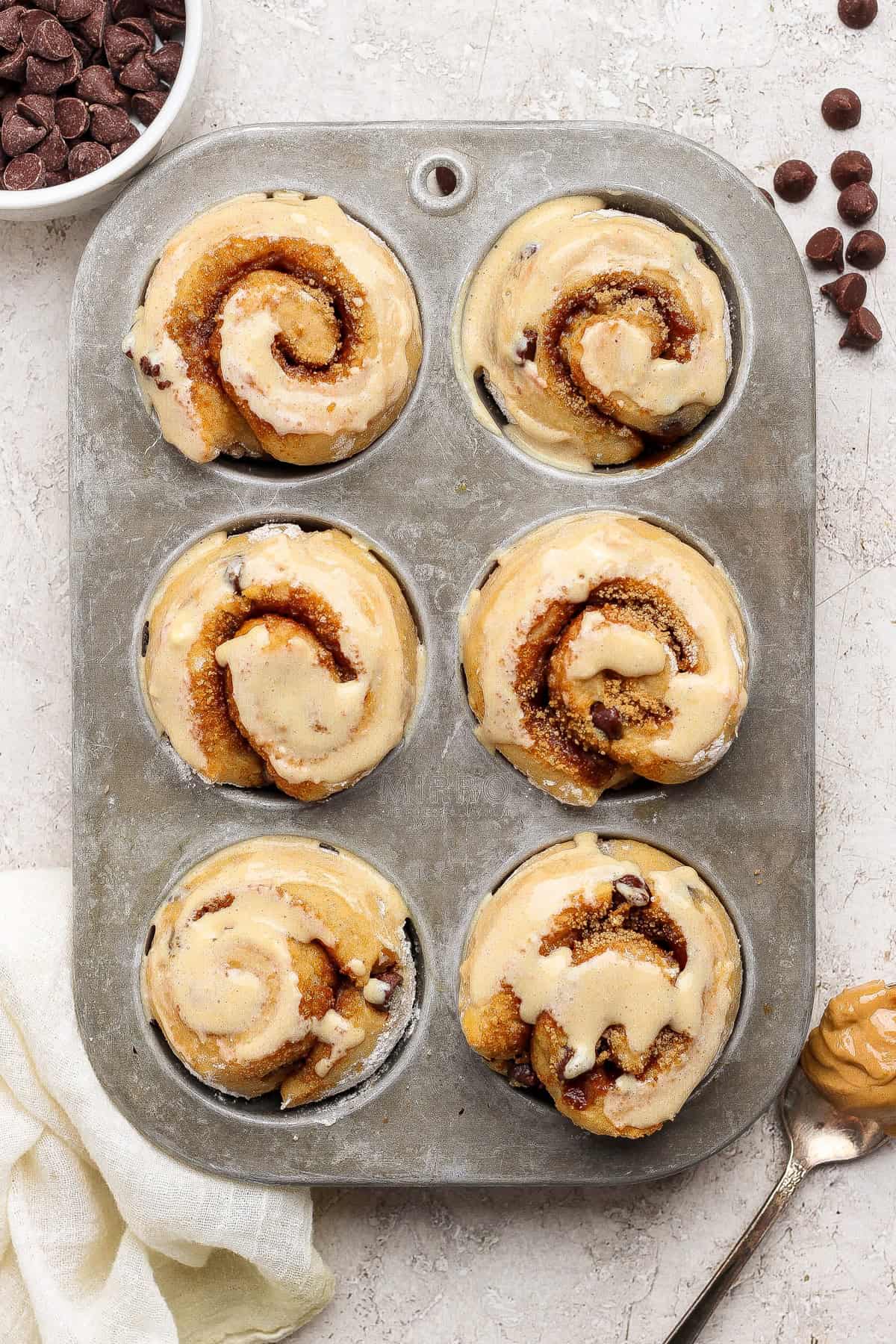 Six unbaked cinnamon rolls with chocolate chips in a metal muffin tin, with a bowl of chocolate chips and a spoon with peanut butter nearby.
