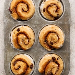Six unbaked cinnamon rolls with chocolate chips in a metal muffin tin, set on a light textured surface.