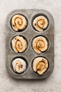 Six unbaked cinnamon rolls with chocolate chips arranged in a metal muffin tin on a textured surface.