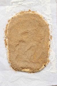 A sheet of dough rolled out on parchment paper and covered evenly with a layer of brown sugar and cinnamon mixture, with some flour scattered around the edges.