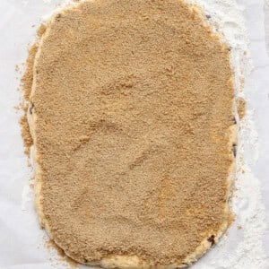 A sheet of dough rolled out on parchment paper and covered evenly with a layer of brown sugar and cinnamon mixture, with some flour scattered around the edges.