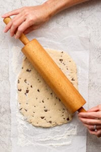 A person rolls out chocolate chip cookie dough on parchment paper using a wooden rolling pin.