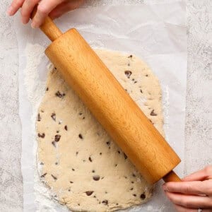 A person rolls out chocolate chip cookie dough on parchment paper using a wooden rolling pin.