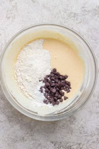 A glass bowl with cookie dough batter, flour, and chocolate chips on a light textured surface.
