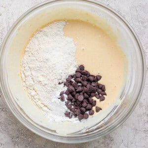 A glass bowl with cookie dough batter, flour, and chocolate chips on a light textured surface.