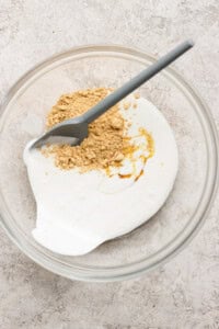 A glass bowl containing yogurt, honey, and ground ginger with a gray spatula on a light textured surface.