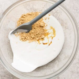 A glass bowl containing yogurt, honey, and ground ginger with a gray spatula on a light textured surface.