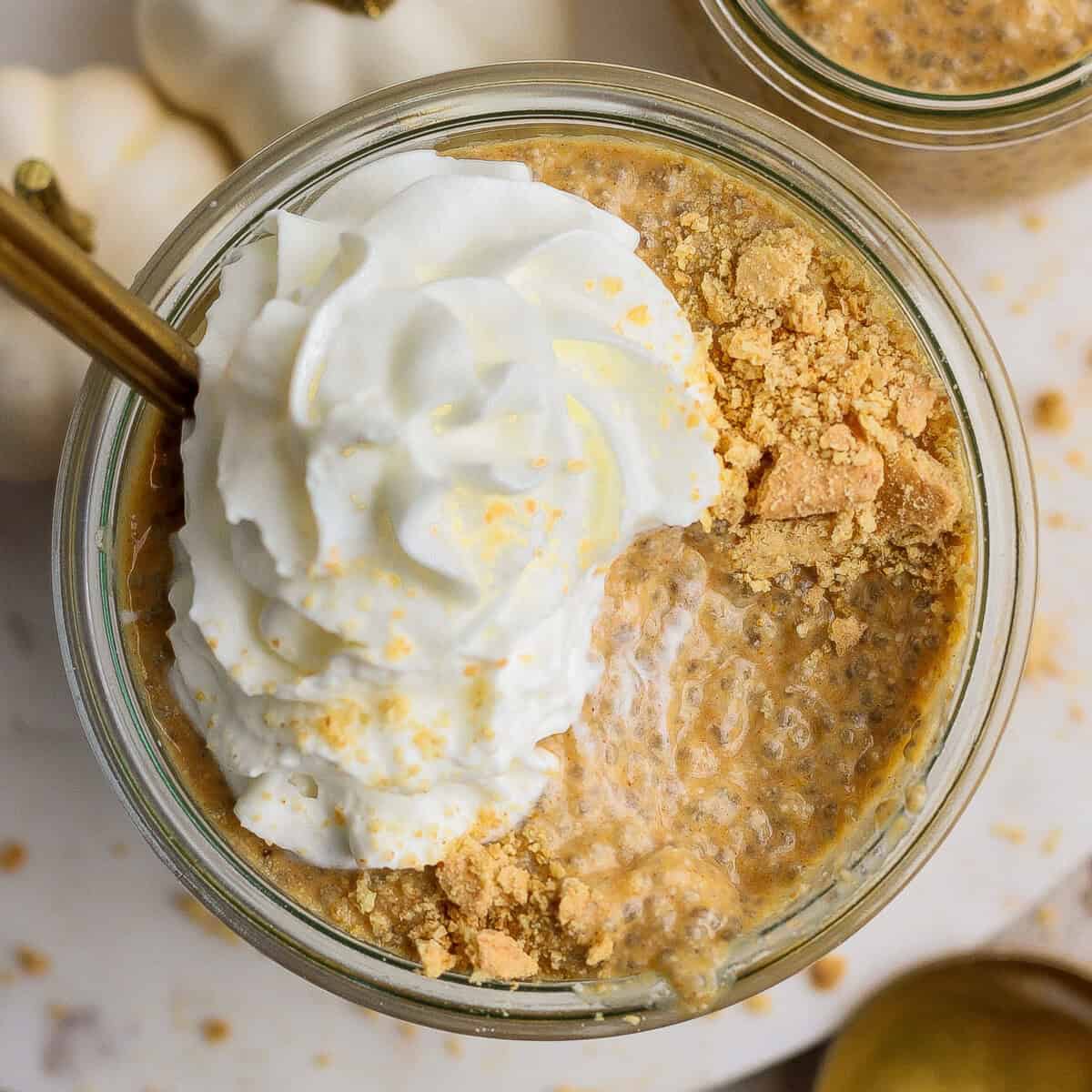 A glass jar filled with pumpkin chia pudding, topped with whipped cream and crushed graham crackers, with a gold spoon inserted.