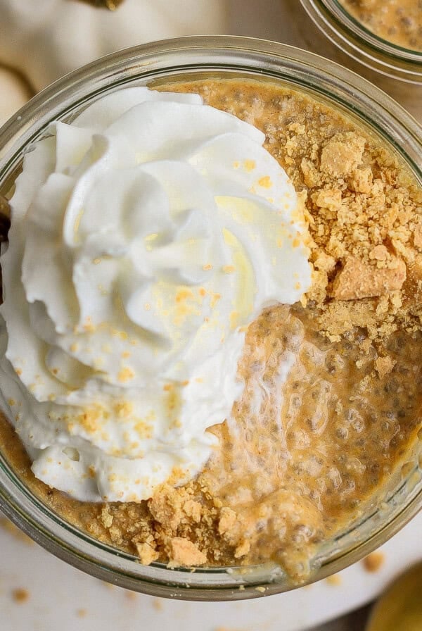 A glass jar filled with pumpkin chia pudding, topped with whipped cream and crushed graham crackers, with a gold spoon inserted.