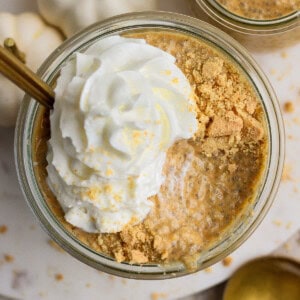 Glass jar filled with pumpkin chia pudding, topped with whipped cream and crushed graham crackers, with a gold spoon and white decorative pumpkins nearby.