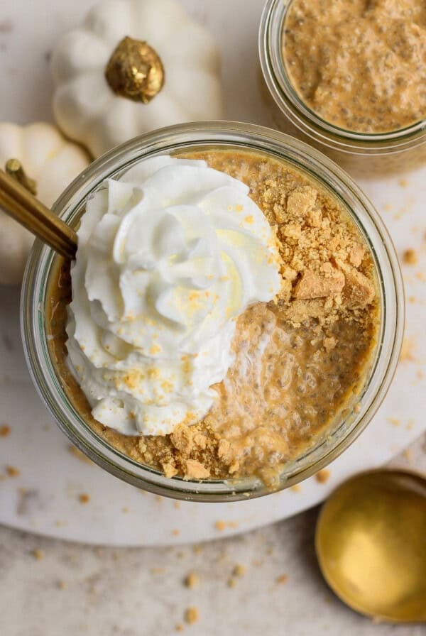 Glass jar filled with pumpkin chia pudding, topped with whipped cream and crushed graham crackers, with a gold spoon and white decorative pumpkins nearby.