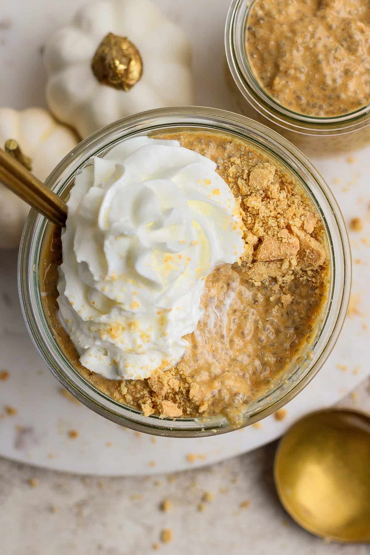 Glass jar filled with pumpkin chia pudding, topped with whipped cream and crushed graham crackers, with a gold spoon and white decorative pumpkins nearby.