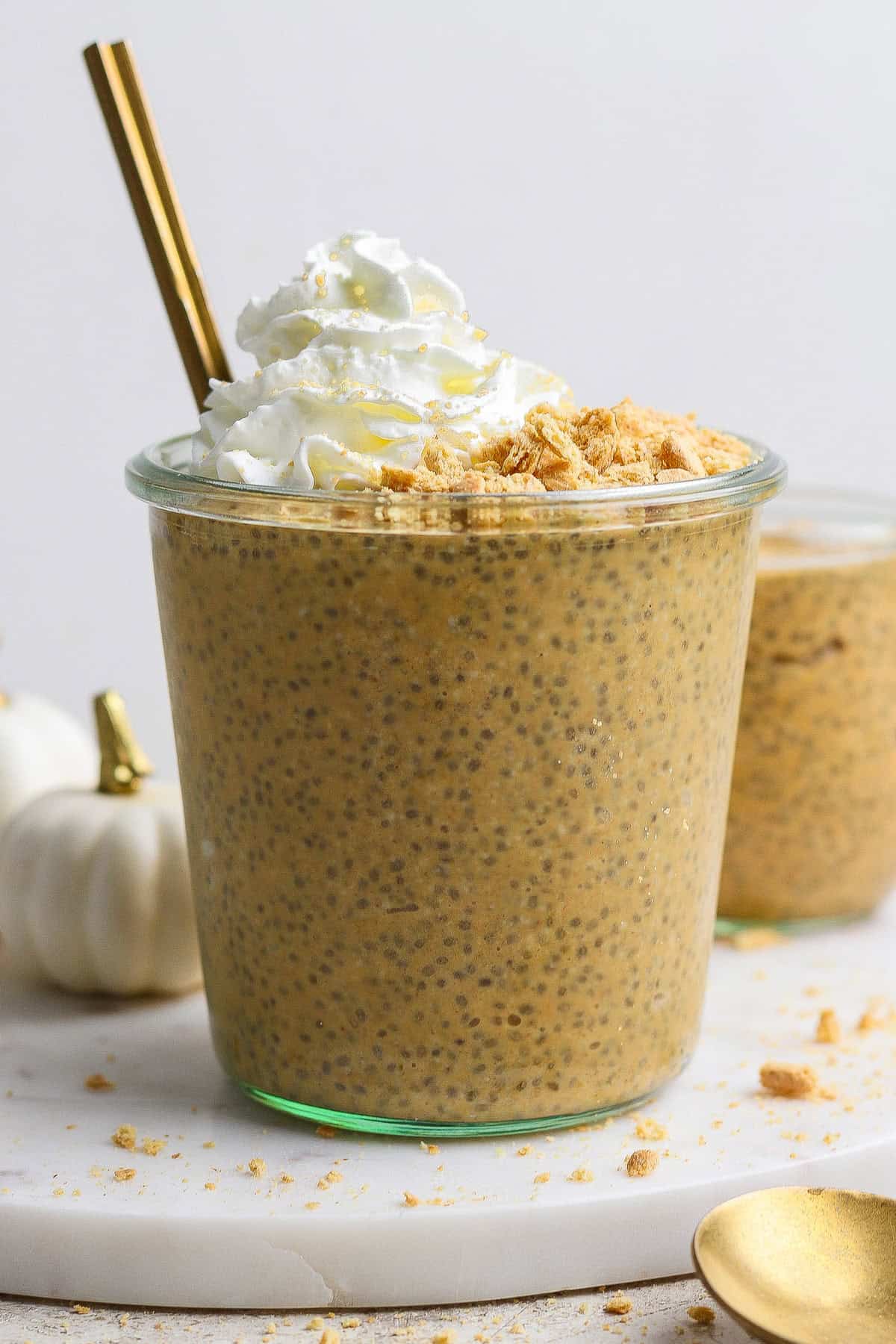 A glass filled with pumpkin chia pudding, topped with whipped cream and crushed crumbs, with a gold spoon and straw, on a white surface.