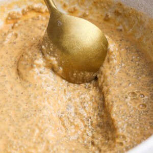 A gold spoon stirs a thick, light brown chia pudding mixture in a white bowl.
