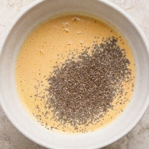 A white bowl containing whisked eggs with a pile of chia seeds on top, set on a light-colored textured surface.