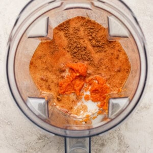Overhead view of a blender containing orange puree, milk, and brown spices on a light-colored surface.