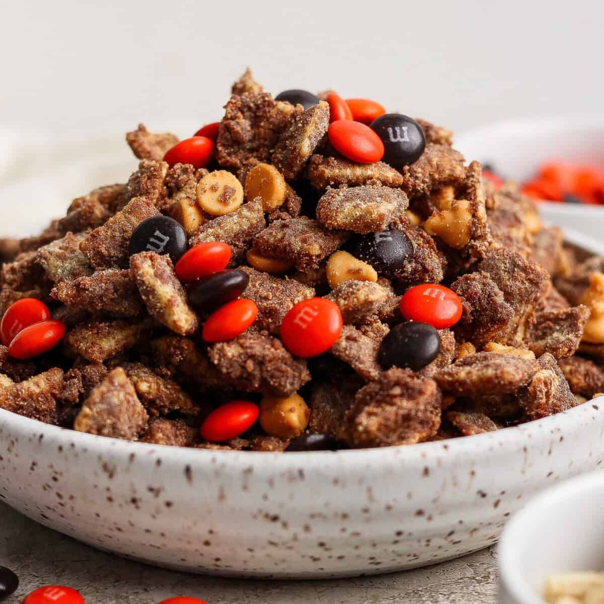 A bowl filled with chocolate-coated nuts or snack mix, topped with orange and brown M&M’s and butterscotch chips.
