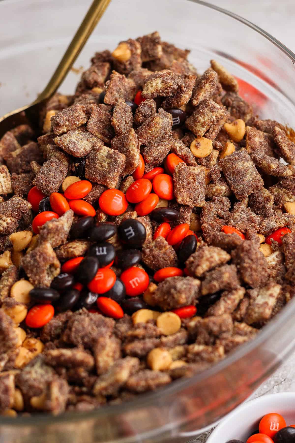 A glass bowl filled with chocolate-coated cereal pieces, peanut butter chips, and red, orange, and brown candy-coated chocolates.