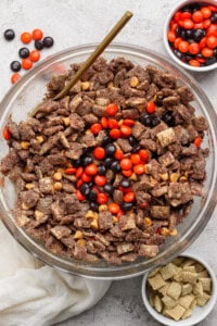 A glass bowl filled with chocolate-coated Chex cereal, peanut butter chips, and orange and brown M&M candies; small bowls with extra M&Ms and plain Chex nearby.