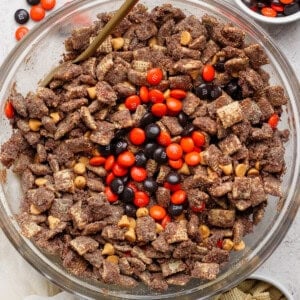 A glass bowl filled with chocolate-coated Chex cereal, peanut butter chips, and orange and brown M&M candies; small bowls with extra M&Ms and plain Chex nearby.