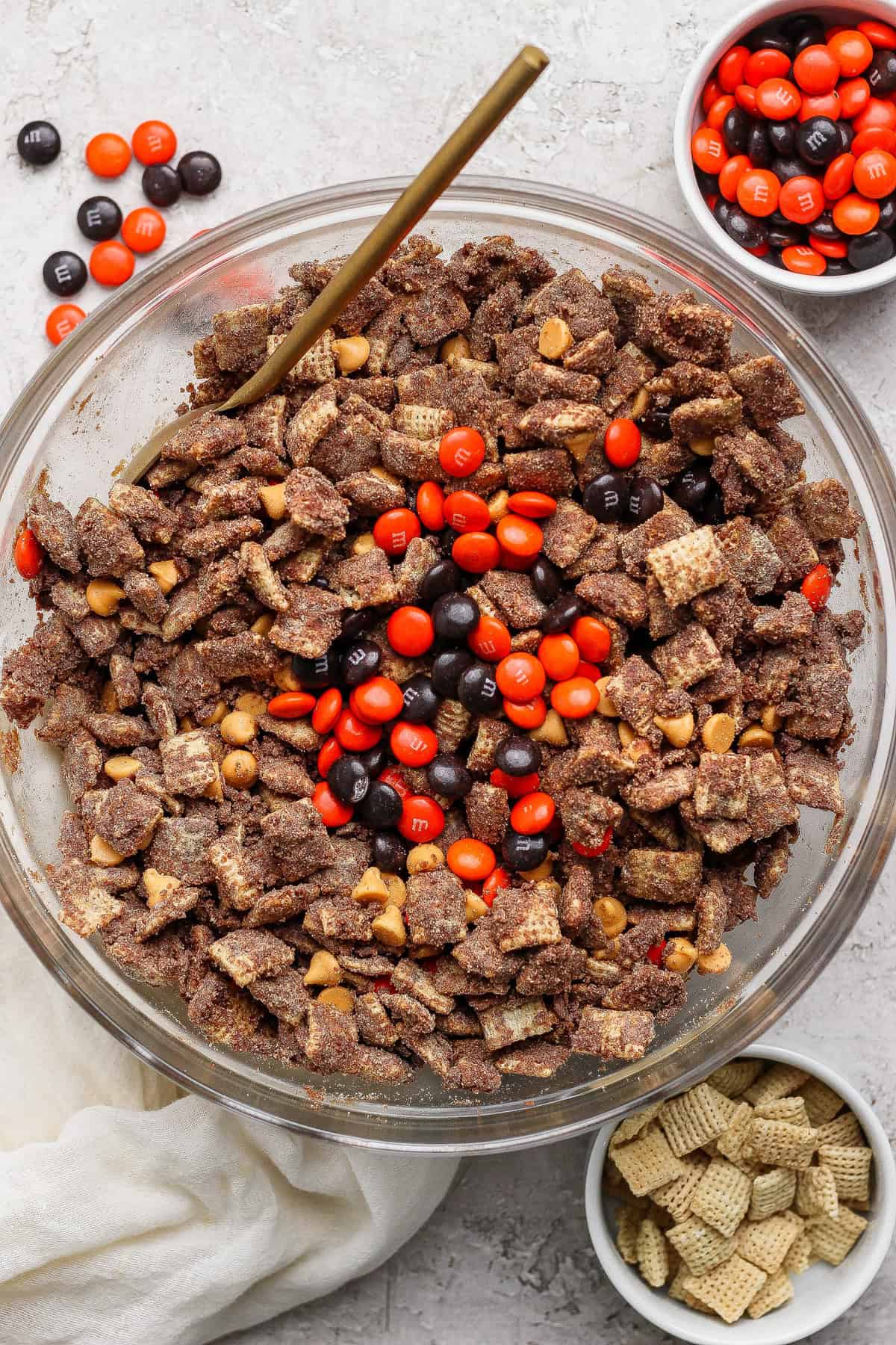 A glass bowl filled with chocolate-coated Chex cereal, peanut butter chips, and orange and brown M&M candies; small bowls with extra M&Ms and plain Chex nearby.