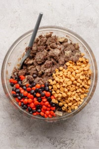 A glass bowl containing chocolate-covered cereal, peanut butter chips, and red and brown candy-coated chocolates, with a gray spoon inside.