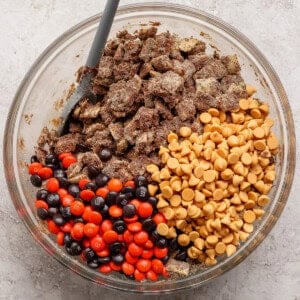 A glass bowl containing chocolate-covered cereal, peanut butter chips, and red and brown candy-coated chocolates, with a gray spoon inside.