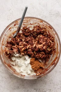 A glass bowl contains chocolate-coated Chex cereal, white powdered sugar, and brown cinnamon, with a gray spatula resting inside.