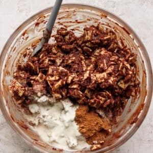A glass bowl contains chocolate-coated Chex cereal, white powdered sugar, and brown cinnamon, with a gray spatula resting inside.