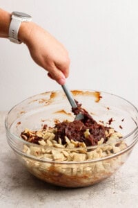 A person mixes chocolate with Chex cereal in a clear glass bowl using a spatula.