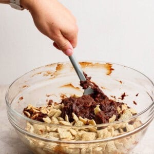 A person mixes chocolate with Chex cereal in a clear glass bowl using a spatula.