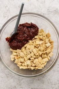 A glass bowl containing Chex cereal and chocolate mixture with a spatula on a light textured surface.