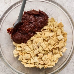 A glass bowl containing Chex cereal and chocolate mixture with a spatula on a light textured surface.