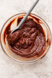 A glass bowl filled with thick chocolate batter and a gray spatula on a light textured surface.
