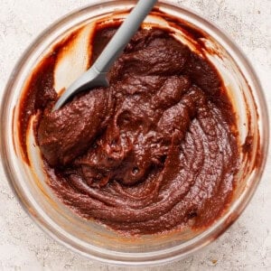 A glass bowl filled with thick chocolate batter and a gray spatula on a light textured surface.