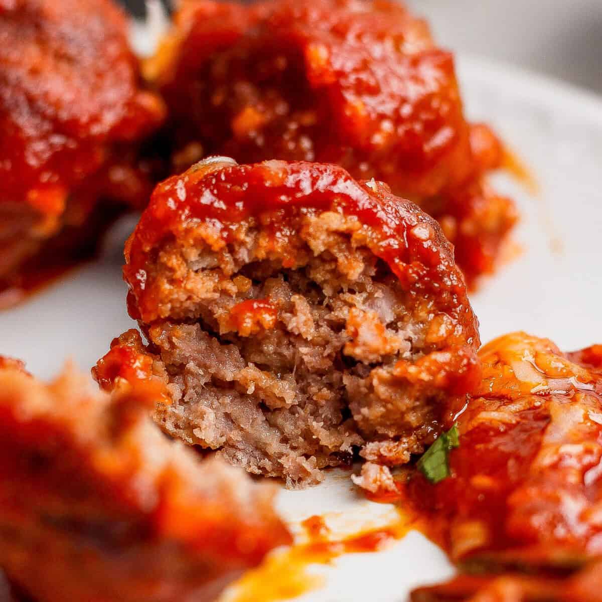 A close-up of a partially eaten meatball covered in tomato sauce, revealing its cooked, ground meat interior.