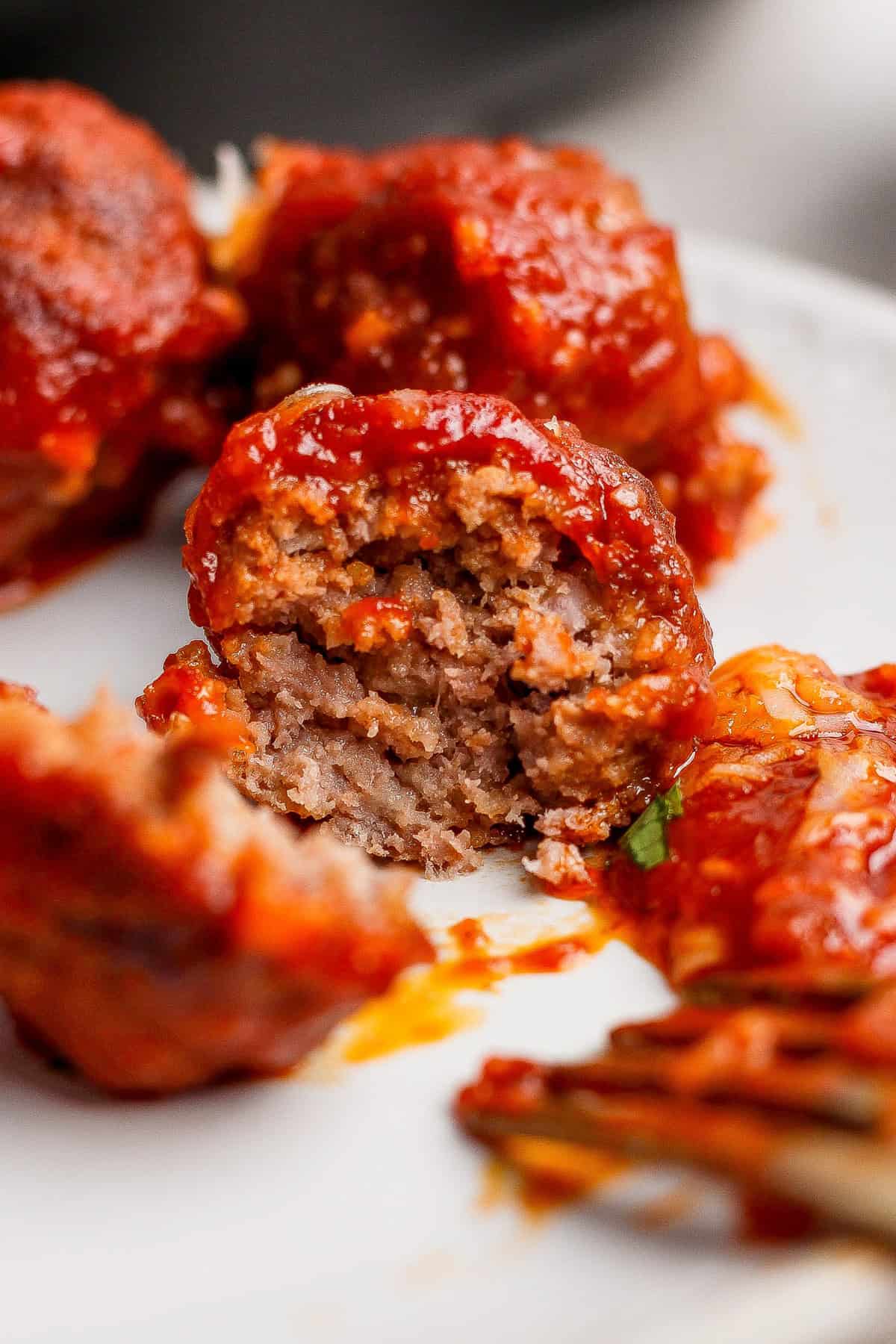 Close-up of meatballs covered in tomato sauce, with one meatball cut open to show the inside texture, on a white plate.