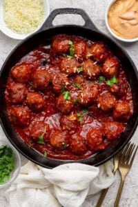 A cast iron skillet filled with meatballs in tomato sauce, garnished with parsley, surrounded by bowls of grated cheese, sauce, and chopped herbs.