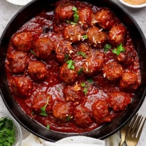 A cast iron skillet filled with meatballs in tomato sauce, garnished with parsley, surrounded by bowls of grated cheese, sauce, and chopped herbs.