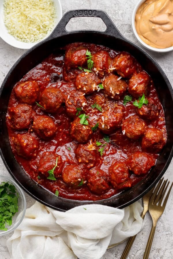 A cast iron skillet filled with meatballs in tomato sauce, garnished with parsley, surrounded by bowls of grated cheese, sauce, and chopped herbs.