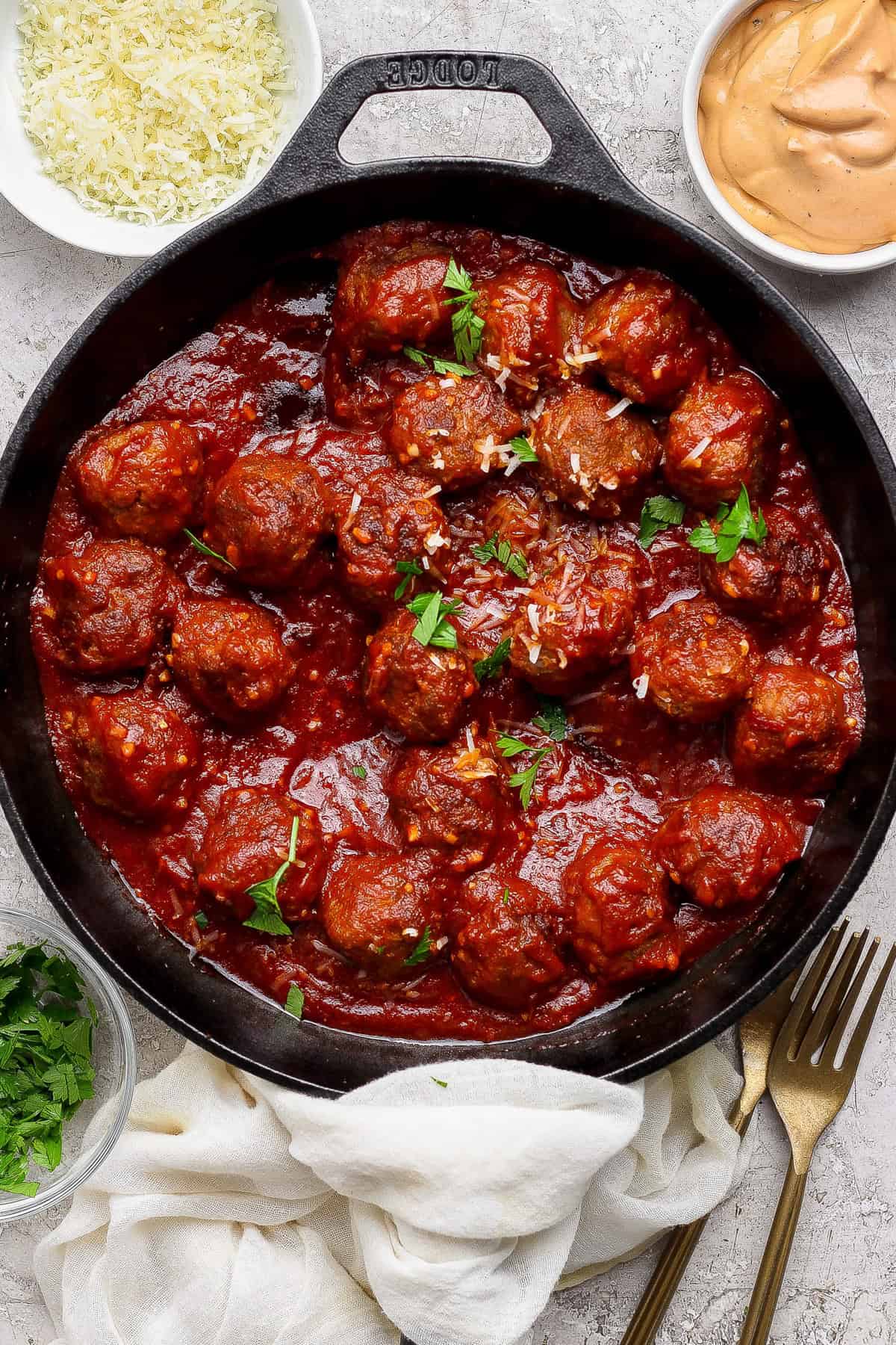 A cast iron skillet filled with meatballs in tomato sauce, garnished with parsley, surrounded by bowls of grated cheese, sauce, and chopped herbs.