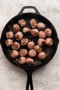 A black cast iron skillet with several beef meatballs being cooked, some browned and some still pink.
