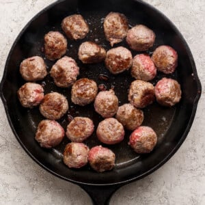 A black cast iron skillet with several beef meatballs being cooked, some browned and some still pink.