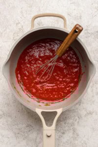 A saucepan filled with red tomato sauce and a metal whisk with a wooden handle resting inside, placed on a light-colored surface.
