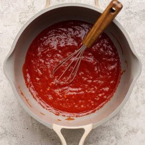 A saucepan filled with red tomato sauce and a metal whisk with a wooden handle resting inside, placed on a light-colored surface.