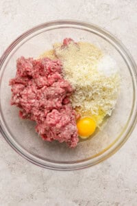 A glass bowl containing raw ground meat, grated cheese, breadcrumbs, and a whole egg on a light-colored surface.