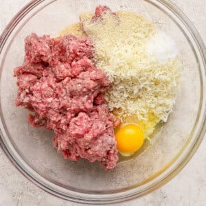 A glass bowl containing raw ground meat, grated cheese, breadcrumbs, and a whole egg on a light-colored surface.