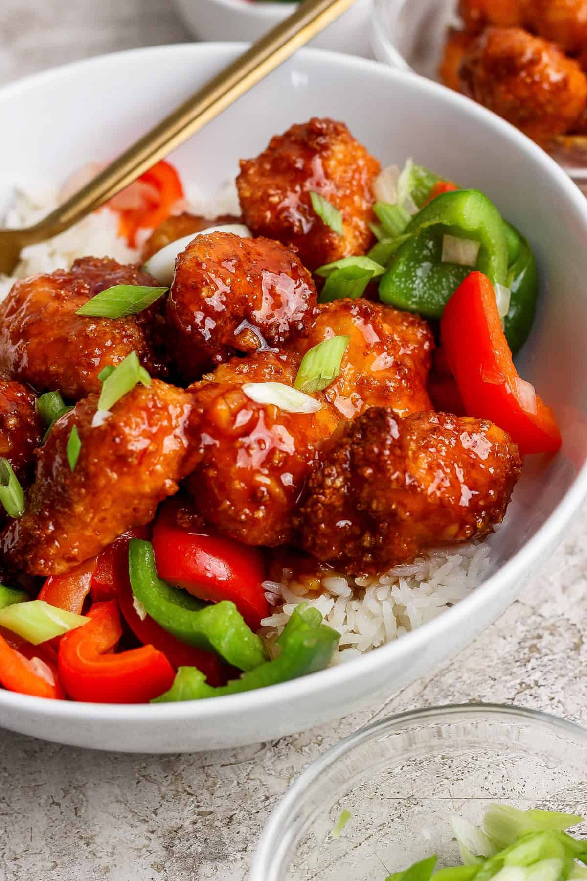 A bowl of white rice topped with crispy glazed chicken pieces, sliced red and green bell peppers, and chopped green onions.