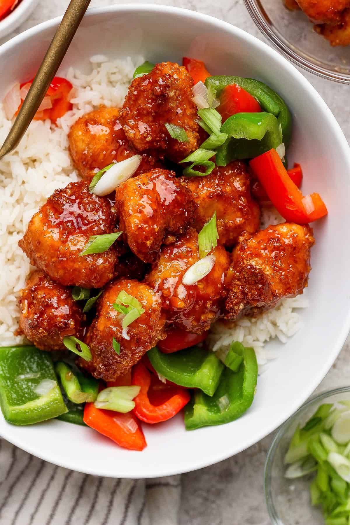 A bowl of white rice topped with glazed chicken pieces, chopped green onions, and sliced red and green bell peppers.