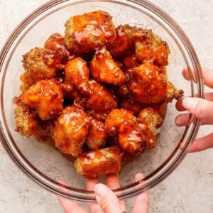 A clear glass bowl filled with breaded, sauce-coated cauliflower bites is being held by two hands over a light-colored surface.